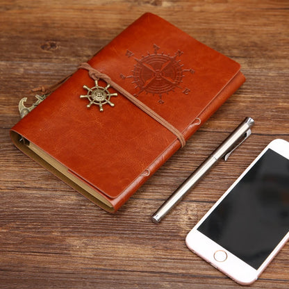 Brown leather notebook with ship wheel design, pen, and phone on wooden surface