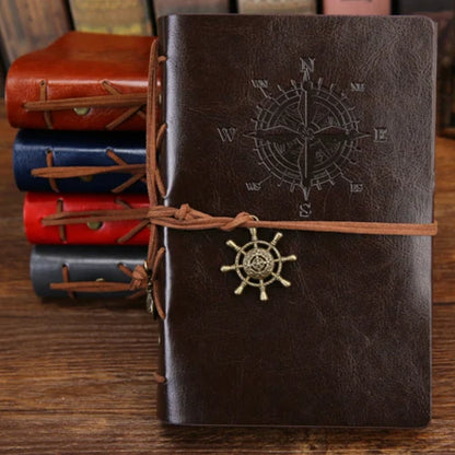 Brown leather notebook with compass and ship wheel design, tied with brown string, on a wooden surface with other notebooks in the background.