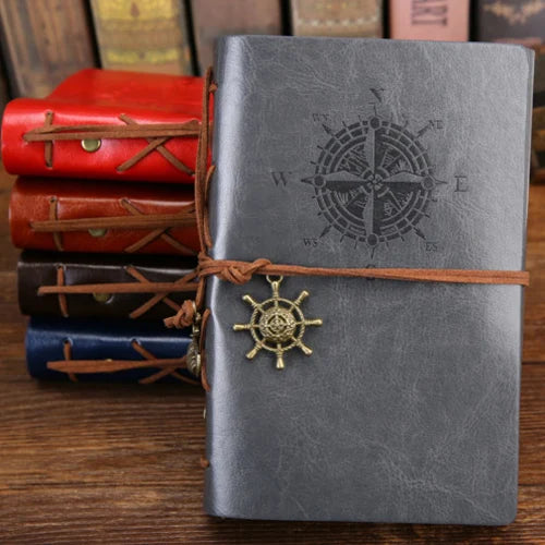 Gray notebook with compass design and leather strap, surrounded by other notebooks on a wooden surface.