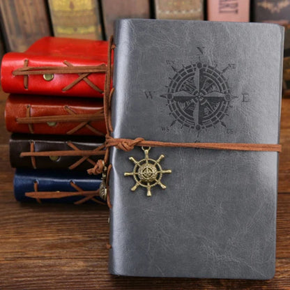 Gray notebook with compass design and leather strap, surrounded by other notebooks on a wooden surface.